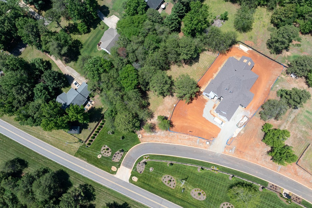 Aerial view of a home under construction with exposed red clay lot, curved driveway, and surrounding trees