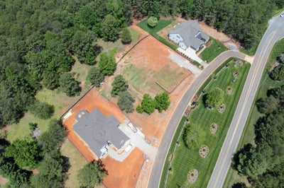 Aerial view of two homes on large lots, one under construction with exposed red clay soil, beside a curved road.