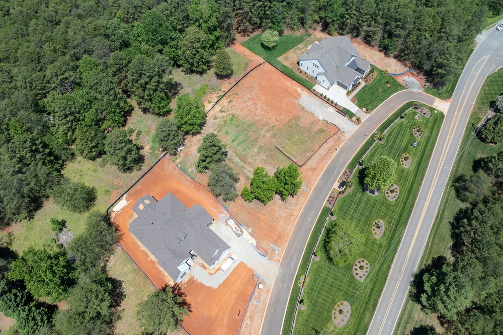 Aerial view of two homes on large lots, one under construction with exposed red clay soil, beside a curved road.