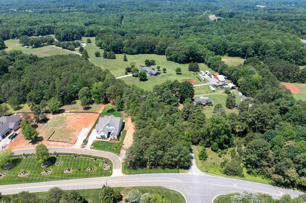 Aerial view of a new home on a large lot surrounded by trees, open land, and a rural neighborhood road.