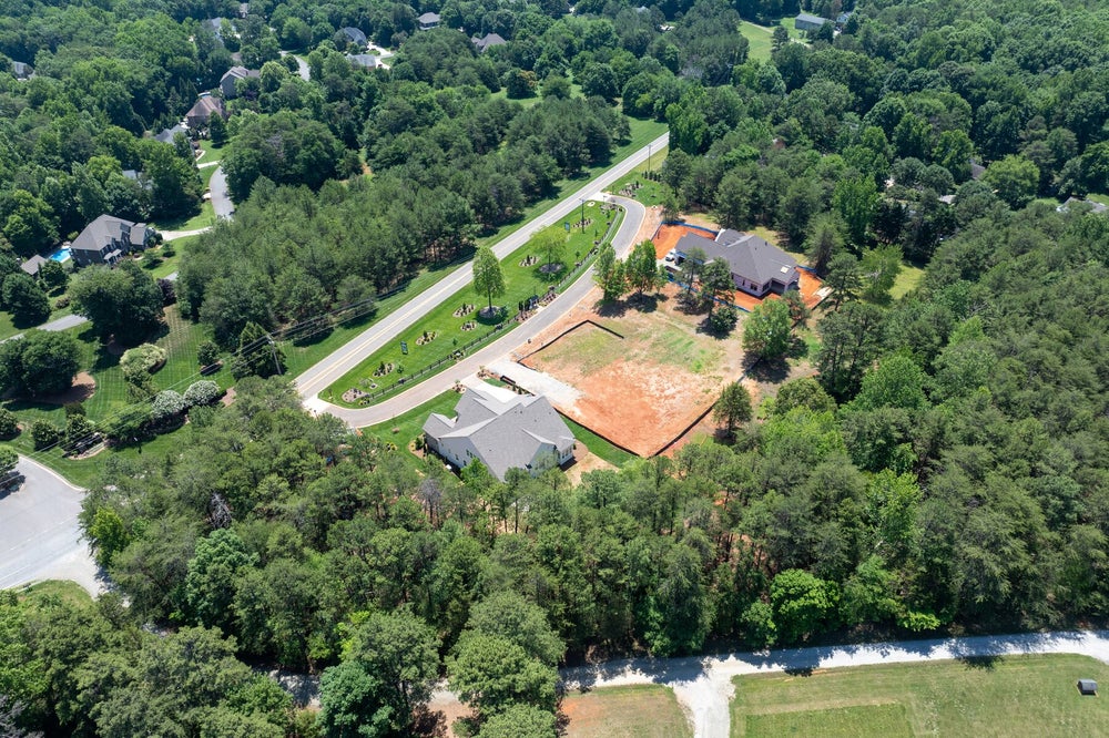 Aerial view of a residential subdivision with two homes, cleared lots under construction, and tree-lined streets surrounded by wooded neighborhood.