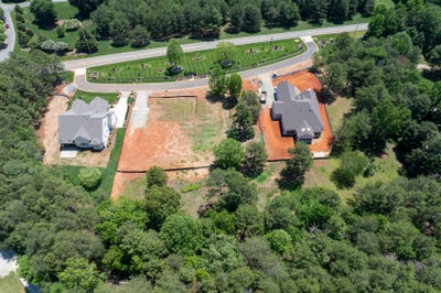 Aerial view of two new homes under construction on large wooded lots near a curved neighborhood entrance road.