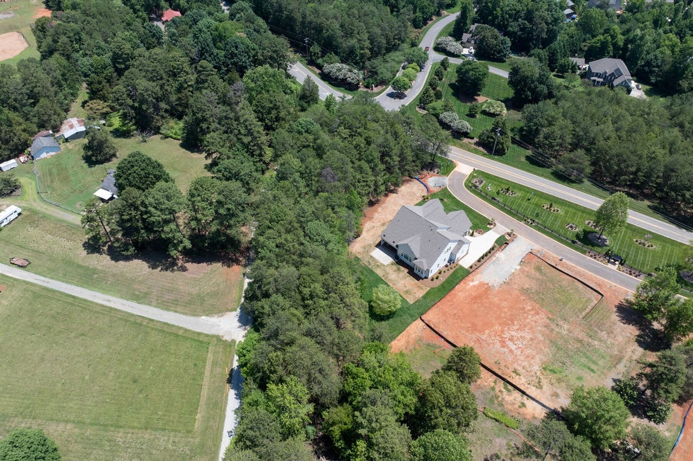Aerial view of a new white home on a wooded lot with cleared land and a curved driveway nearby.