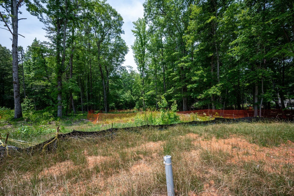 Vacant wooded lot with orange construction fencing, silt barrier, and utility pipe stub at ground level.