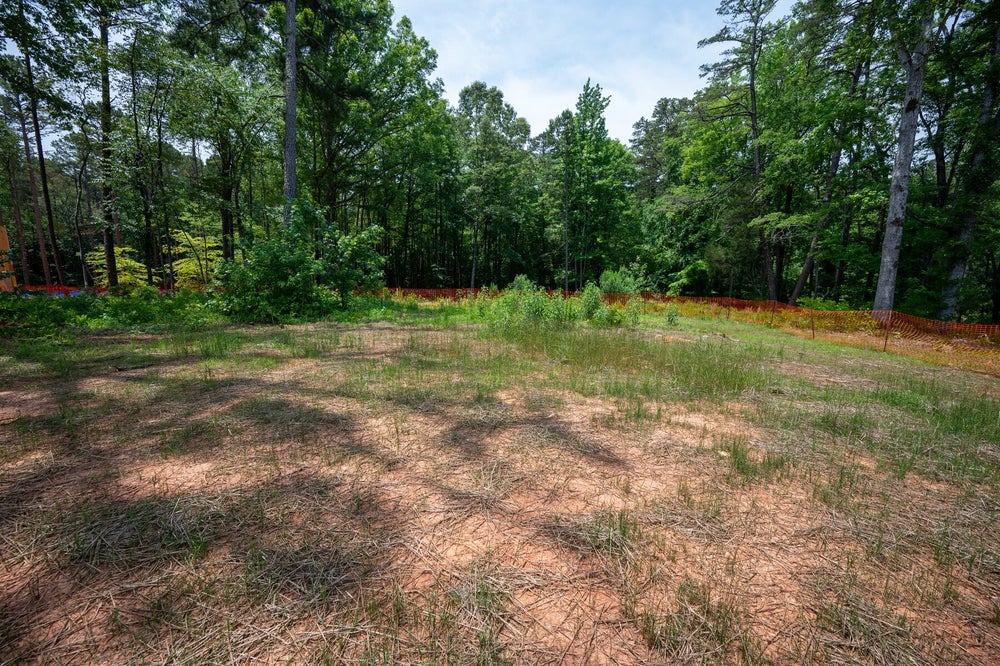 Vacant wooded lot with cleared dirt ground, sparse grass, and orange construction fencing along tree line