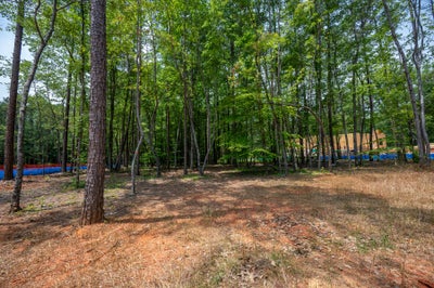 Wooded residential lot with tall trees, dirt ground, and a home under construction visible in the background
