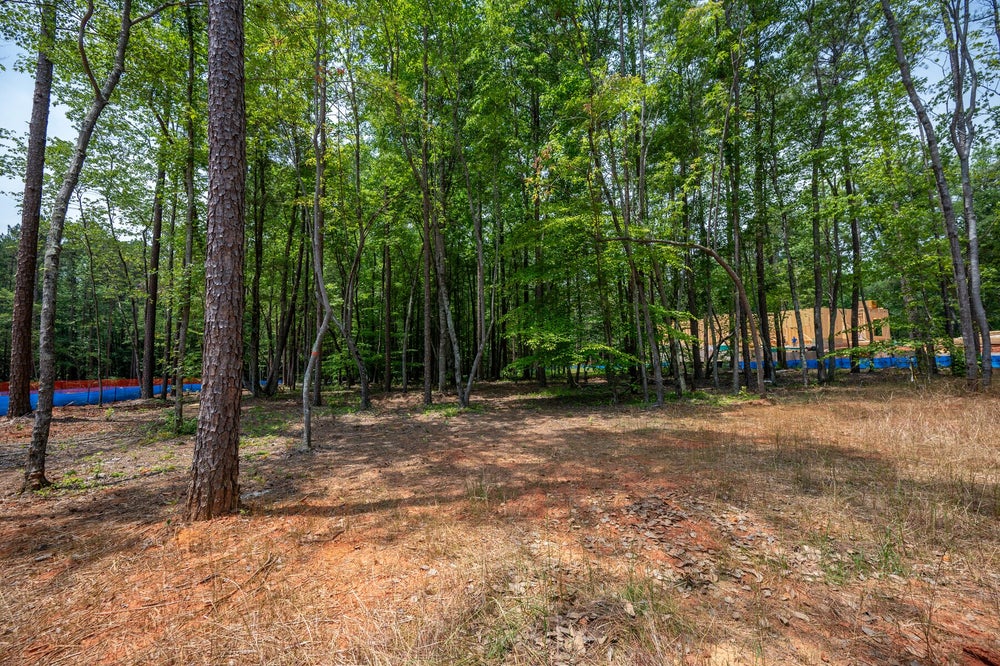 Wooded residential lot with tall trees, dirt ground, and a home under construction visible in the background