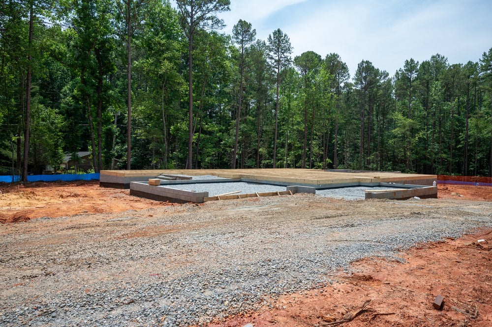 New home construction site showing concrete foundation and wood subfloor framing surrounded by trees