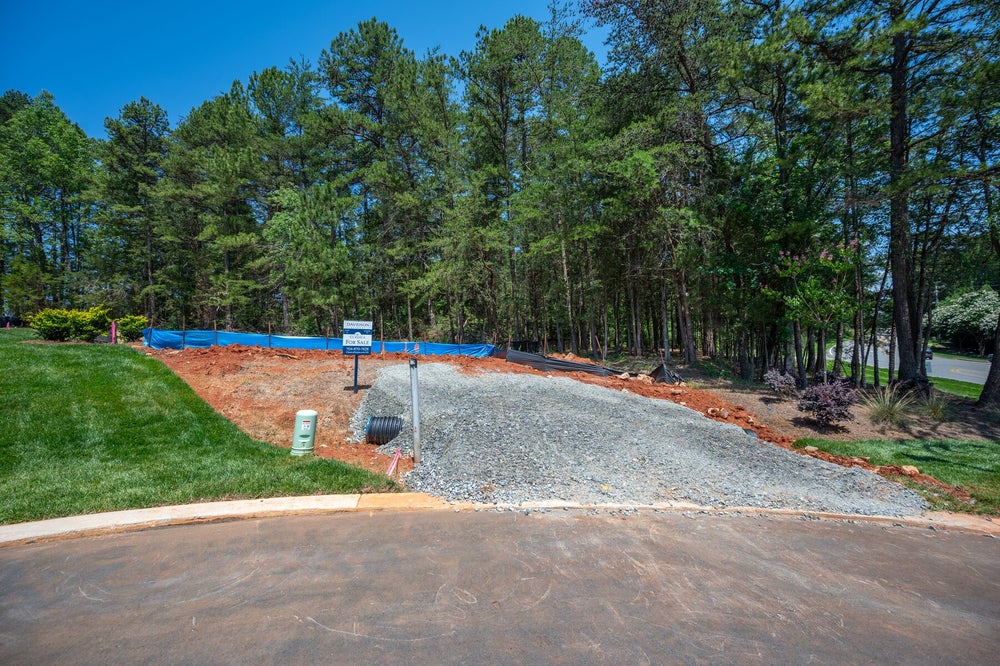 Vacant residential lot for sale with gravel driveway entrance, red clay soil, and pine trees in the background.
