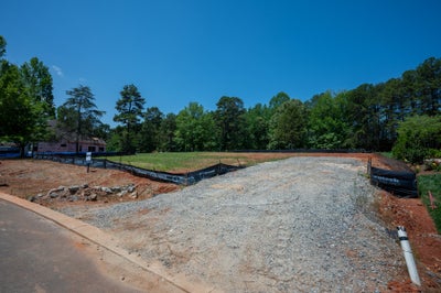 Cleared residential lot with gravel driveway entrance, erosion control fencing, and trees in background