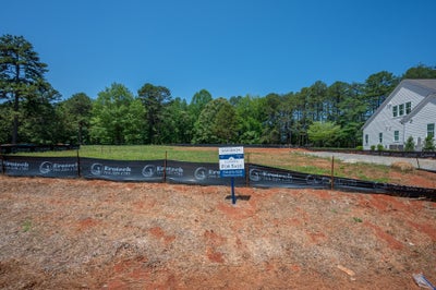 Vacant lot for sale with silt fence, red clay soil, and trees, next to a white home under construction.