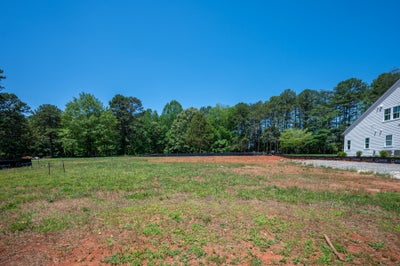 Empty residential lot with cleared dirt and grass, wooded tree line, and white home visible on right side