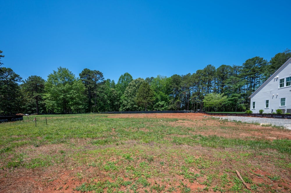 Empty residential lot with cleared dirt and grass, wooded tree line, and white home visible on right side
