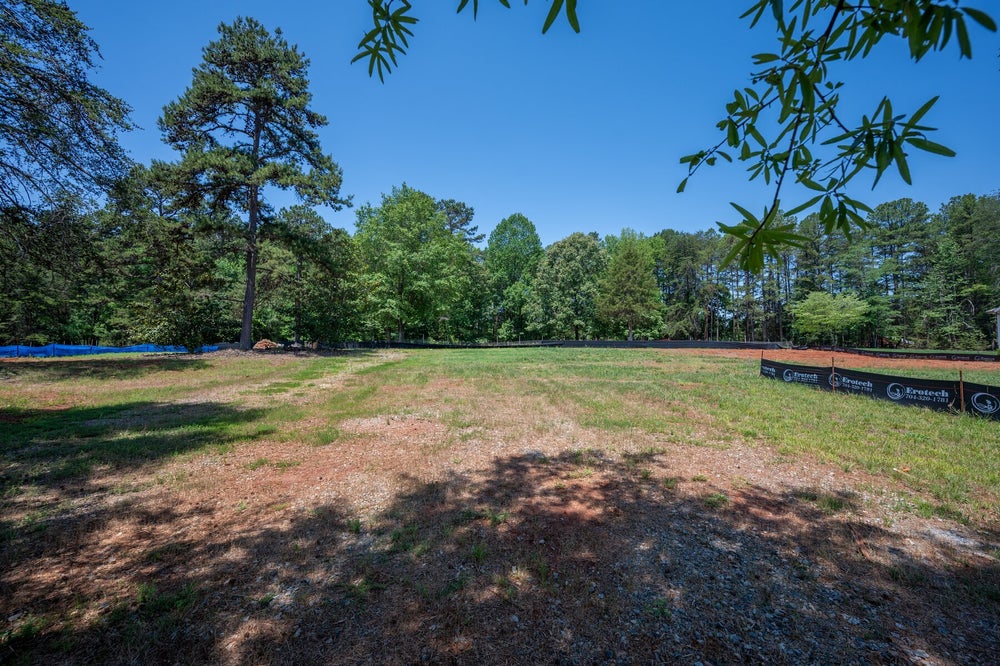 Empty residential lot with cleared land, erosion control fencing, and wooded tree line in background