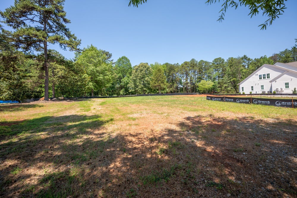 Empty residential lot with grassy yard, construction fencing, and white home visible at edge of wooded tree line