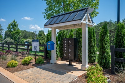 Covered mailbox kiosk with white pergola, metal roof, and cluster mailboxes on brick paver pad near community entrance