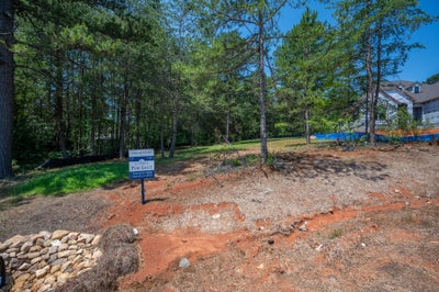 Vacant lot for sale with red clay soil, pine trees, and a neighboring home under construction nearby.