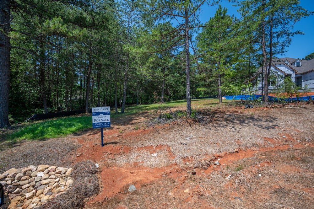 Vacant lot for sale with red clay soil, pine trees, and a neighboring home under construction nearby.