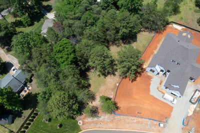 Aerial view of a residential lot with cleared red clay soil next to a home under construction, surrounded by trees.