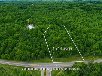 Aerial view of a 1.714-acre wooded lot with marked boundaries along a road, surrounded by trees and a small pond nearby.