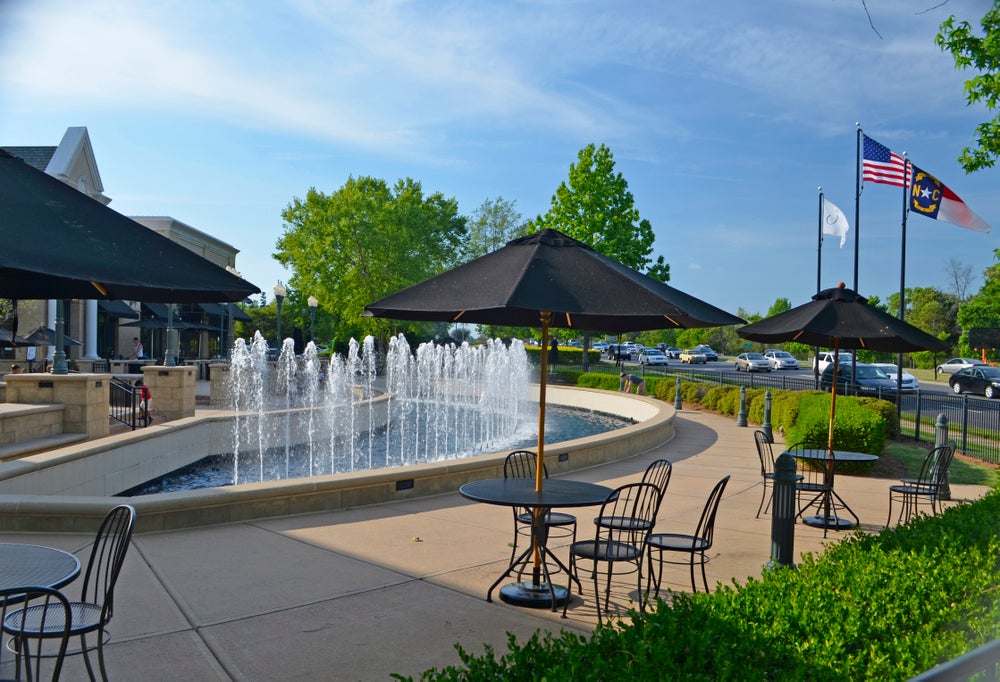 Outdoor plaza with large water fountain, patio tables with umbrellas, and US and North Carolina flags nearby