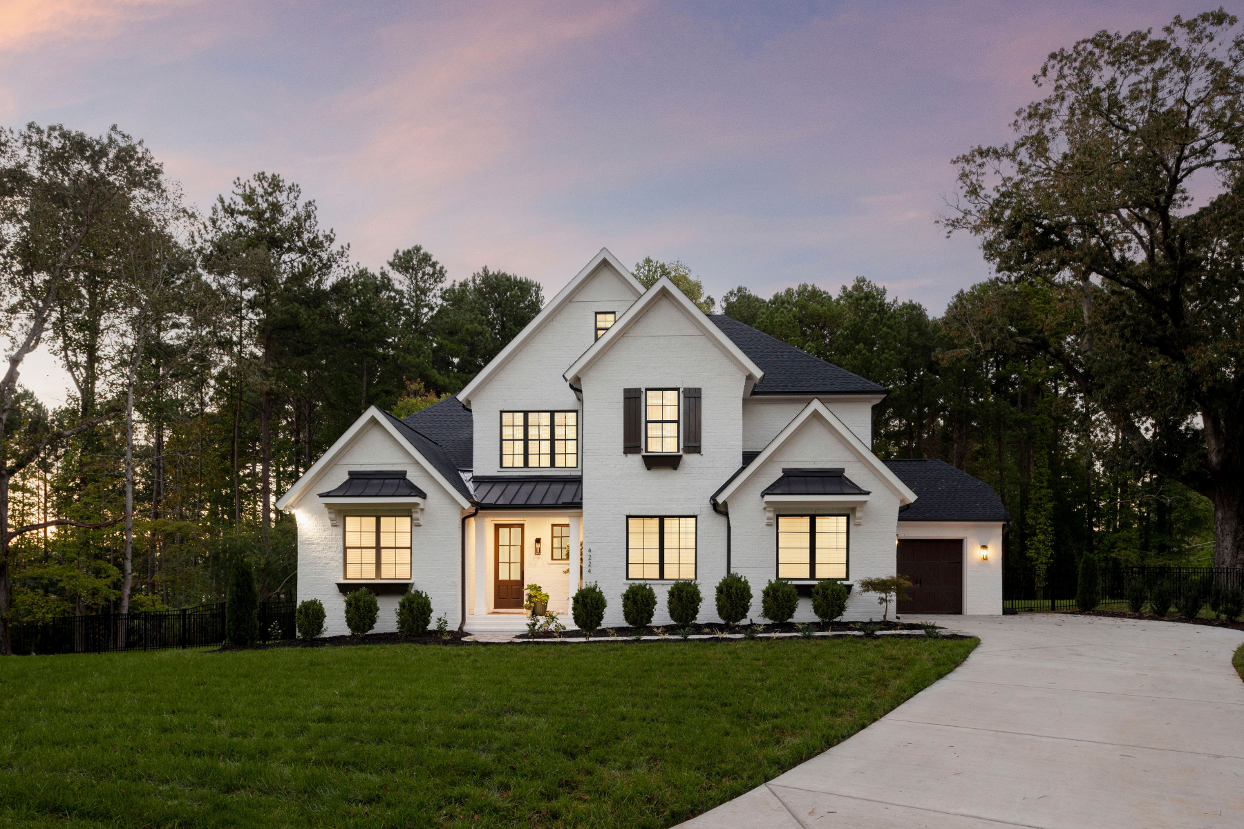 Two-story white brick home with dark roof, black shutters, attached garage, and curved driveway at dusk