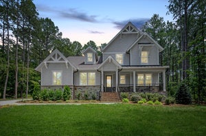 Two-story craftsman home with gray shingle siding, stone base, covered front porch, and green lawn surrounded by trees