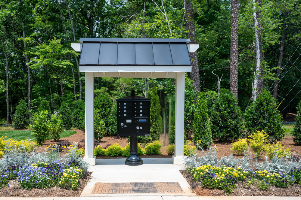 Community mailbox kiosk with black metal roof, white posts, and multi-unit mail cluster box surrounded by landscaped flower beds.