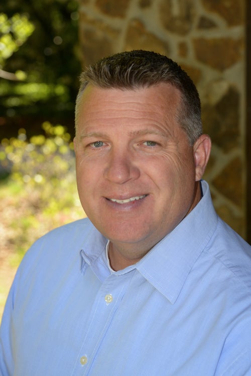 Professional headshot of a smiling middle-aged man in a light blue button-up shirt outdoors