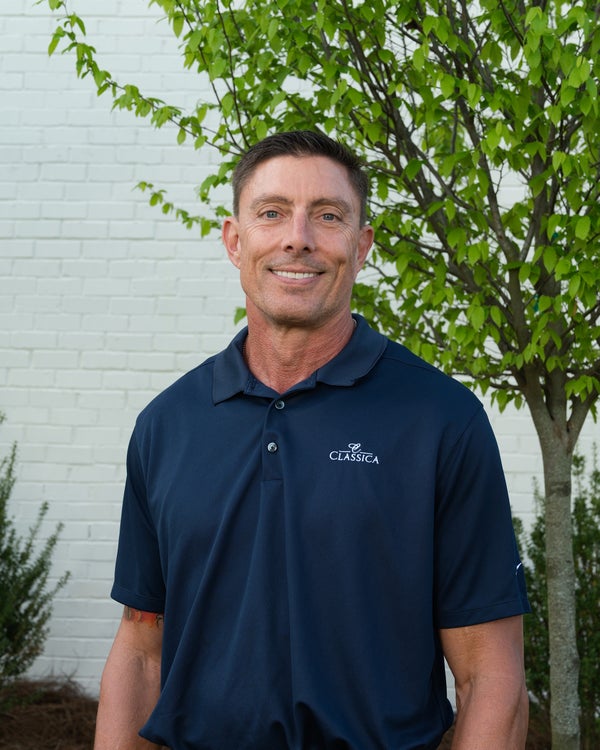 Man wearing a navy Classica polo shirt, smiling outdoors in front of a white brick wall and green tree.
