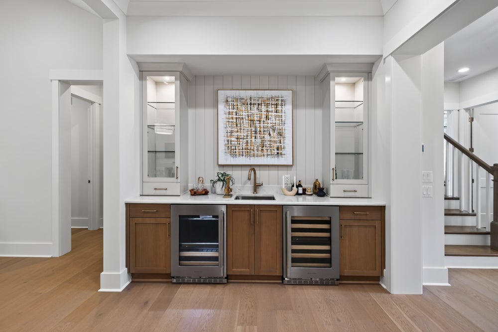 Built-in wet bar with wood cabinets, two beverage fridges, sink, and glass display cabinets on white shiplap wall