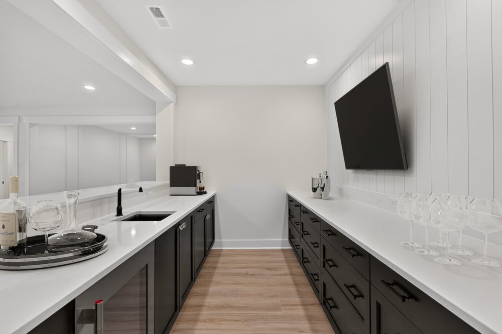 Wet bar with dark cabinets, white countertops, wall-mounted TV, and wine glasses on shiplap accent wall