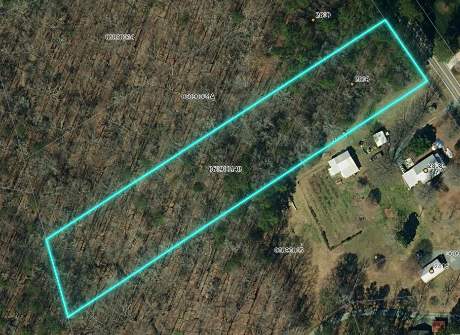 Aerial parcel map showing wooded lot with cyan boundary lines and nearby residential properties along a road.