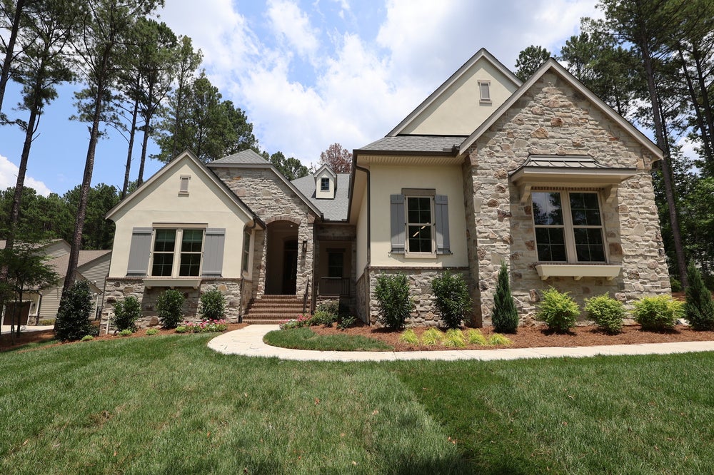 Single-story home with stone and stucco exterior, gray shutters, arched entry, and landscaped front yard surrounded by trees