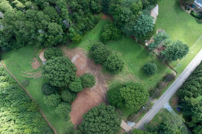 Aerial view of vacant land lot with scattered trees, cleared dirt areas, and a road along the right edge.
