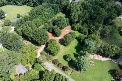 Aerial view of a wooded residential lot with cleared dirt areas, green lawn, and a road along the edge.