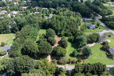 Aerial view of a wooded residential lot with cleared areas, surrounded by neighborhood homes and a road.