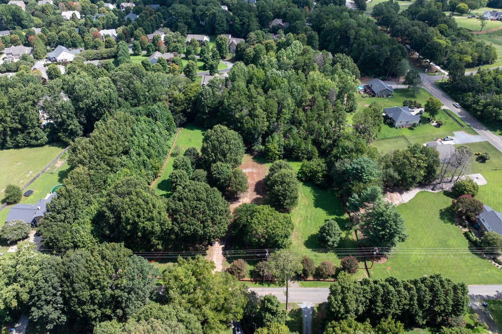 Aerial view of a wooded residential lot with cleared areas, surrounded by neighborhood homes and a road.