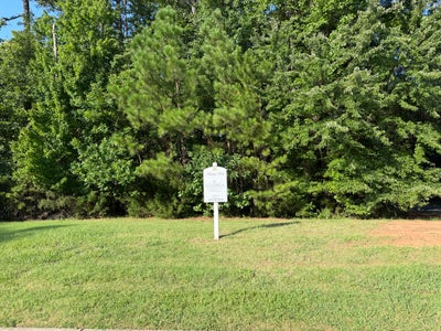 Vacant residential lot with a Classica Homes sign, green grass, and wooded tree line in the background.