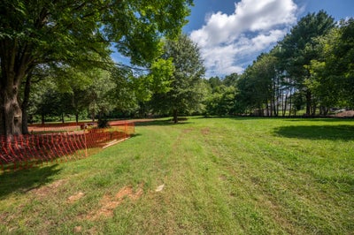 Open grassy lot with mature trees and orange construction fencing marking a build area