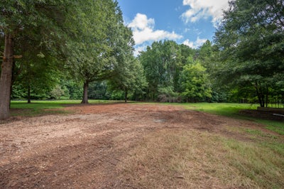 Vacant lot with cleared dirt area, mature trees, and green grass along the edges
