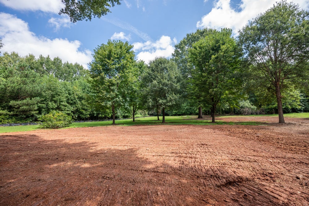 Cleared and graded building lot with red clay soil surrounded by mature trees and green grass