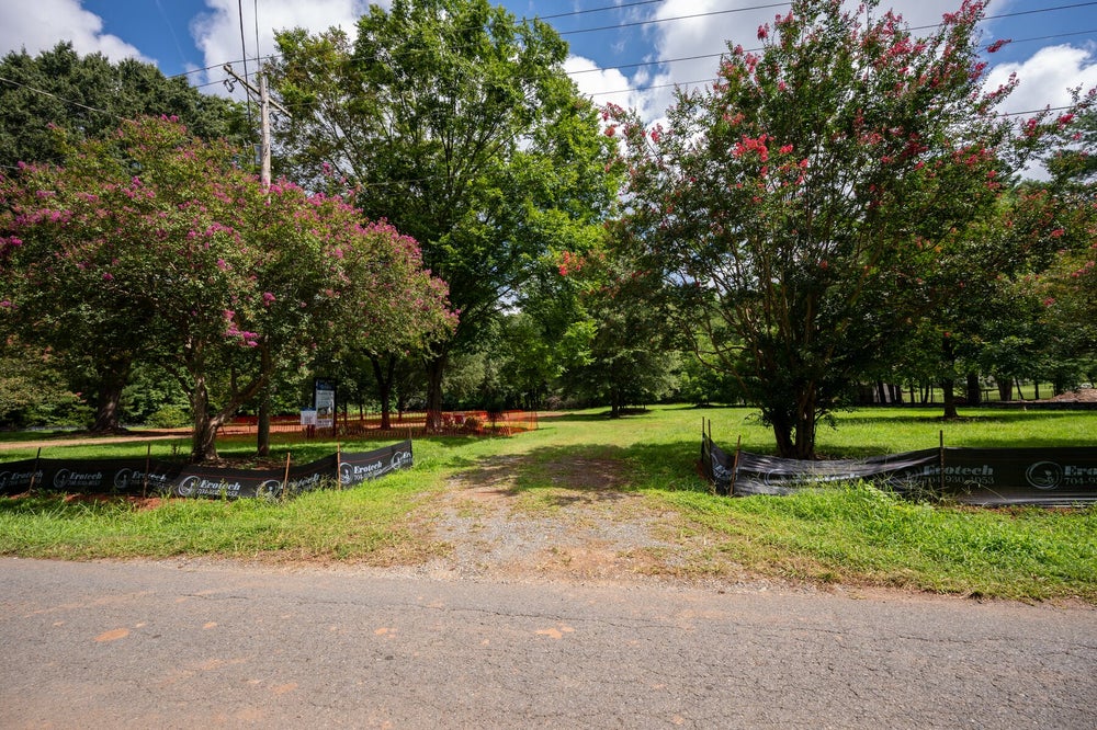 Vacant lot with mature trees, erosion control fencing, and gravel driveway entrance along a road