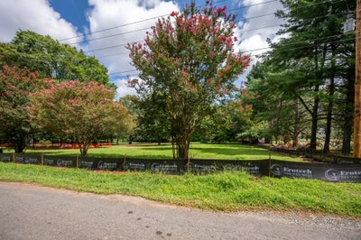 Vacant lot with green grass, blooming crape myrtle trees, and a construction fence along the road