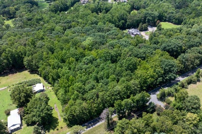 Aerial view of a wooded residential lot bordered by a road, with nearby homes and open green spaces visible.