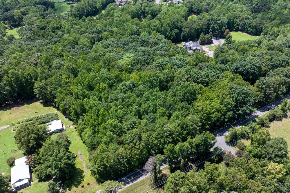 Aerial view of a wooded residential lot bordered by a road, with nearby homes and open green spaces visible.