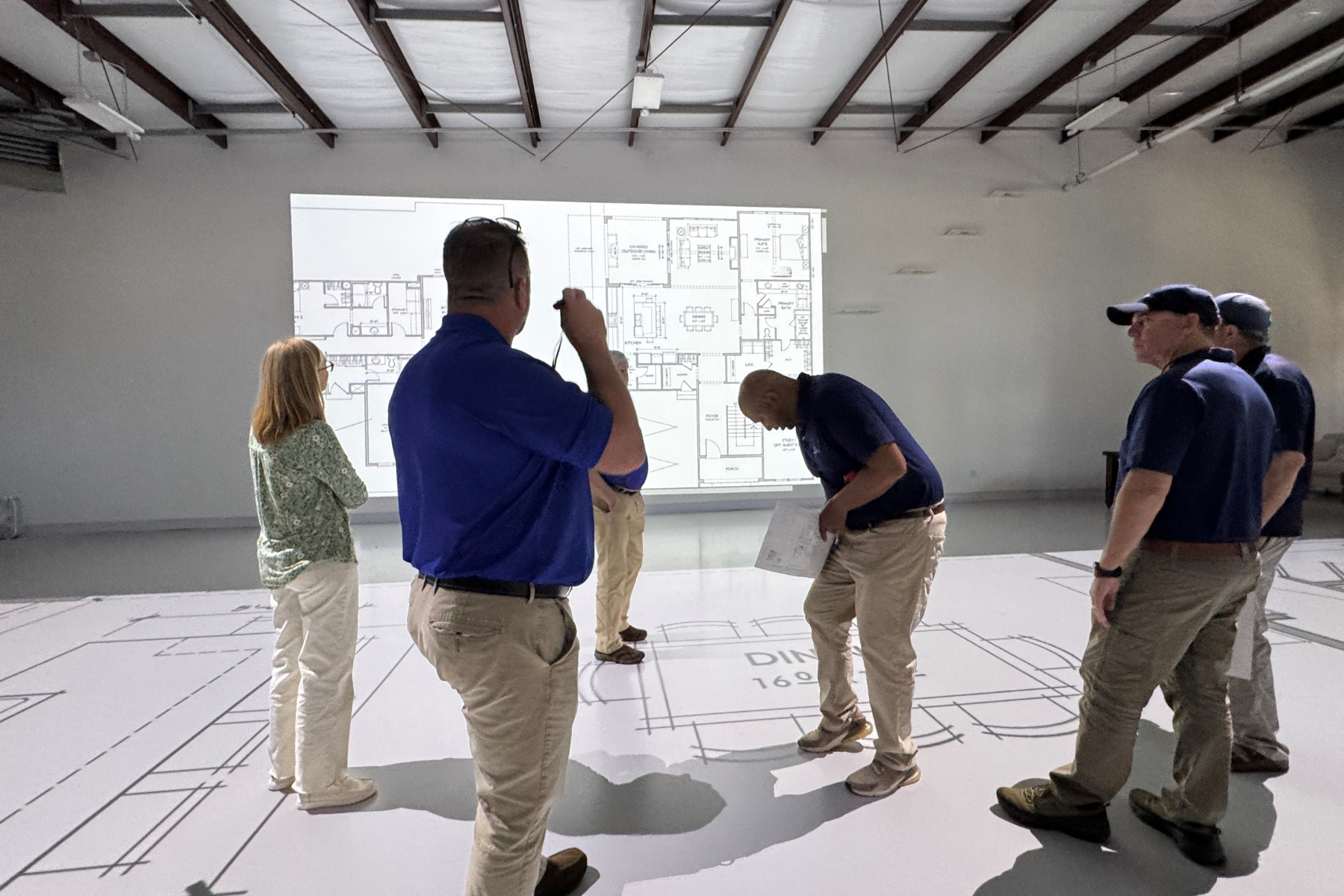 Group of people reviewing a large projected home floor plan in an open warehouse-style room with floor markings.