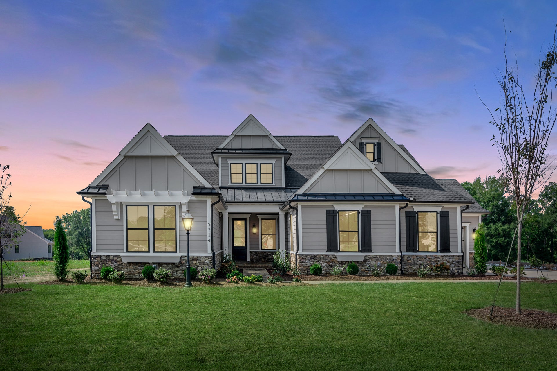 Craftsman-style home with gray board-and-batten siding, stone foundation, covered porch, and large windows at dusk