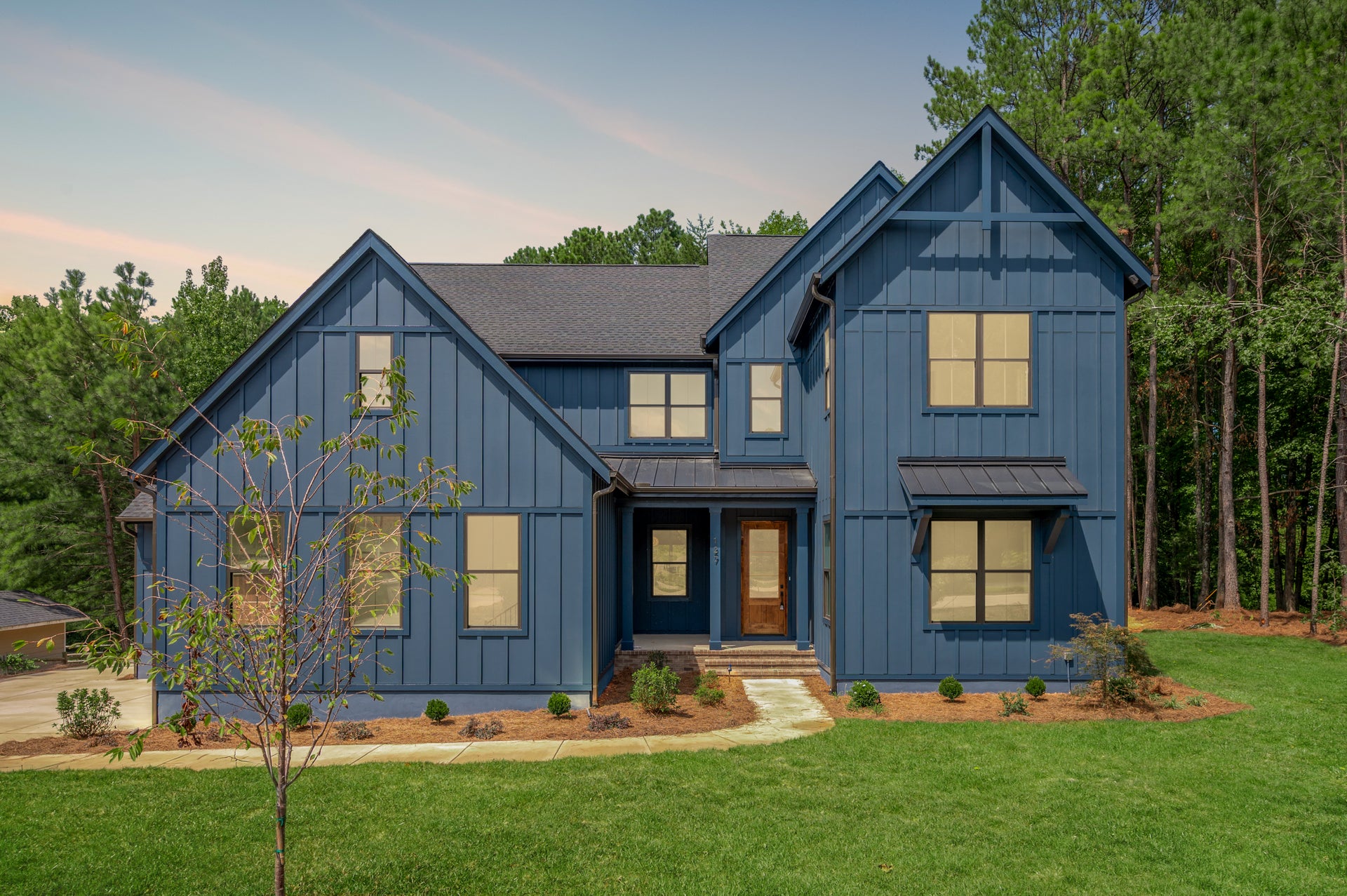 Two-story craftsman home with blue board-and-batten siding, dark roof, and wood front door surrounded by trees