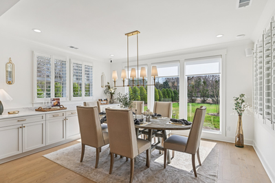 Dining room with round table, six upholstered chairs, gold chandelier, and large windows overlooking a backyard